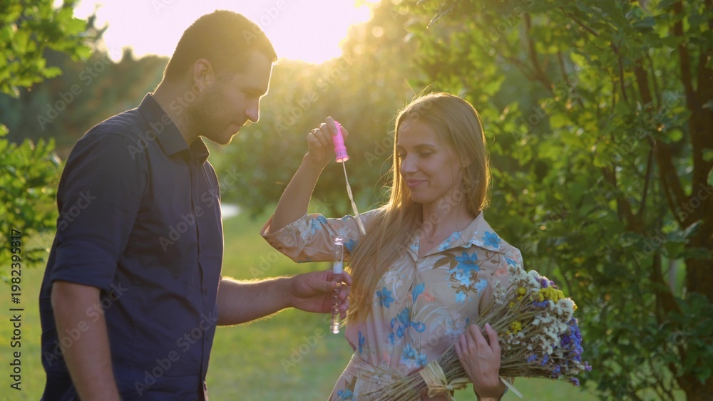 Fototapeta premium Close up of cute couple in love together outside. Man and woman with flowers standing in park and blowing soap bubbles. Love and romantic. Nature. Boyfriend and girlfriend