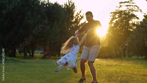 Young Caucasian male parent playing with happy little daughter on nature outdoor. Father spinning girl in park. Kid having fun with dad at sunset. Summertime. Family leisure concept