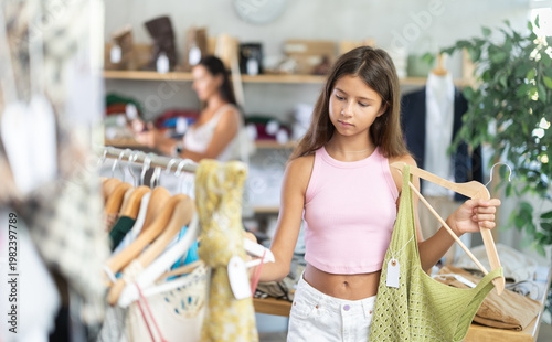 Teenage girl customer choose summer tank top in store, and buyer carefully examines product. Young woman enjoys shopping for stylish summer clothes at modern clothing store