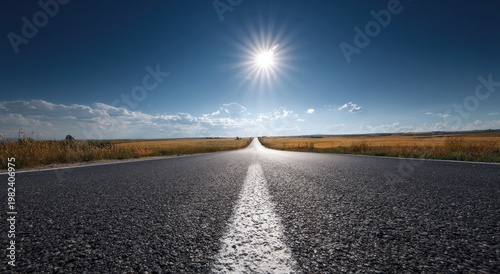 A long, straight asphalt road stretches into the distance under a bright, sunlit sky with scattered clouds over a vast, golden field.