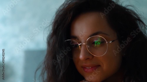 Caucasian young female with long dark hair is wearing glasses, lying on bed and watching movie on her computer. Beautiful woman relaxing and looking at screen with interest. Real-time video, close up.