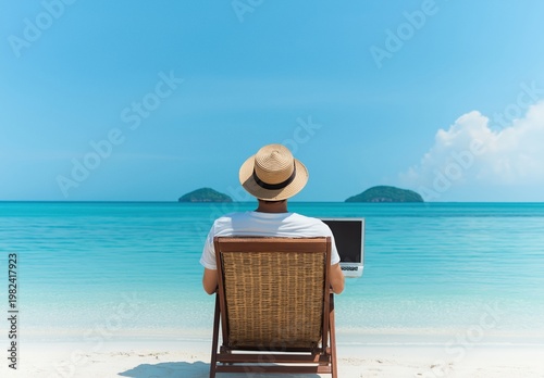 Man wearing hat using laptop, enjoying remote work and tropical vacation on a white sand beach with turquoise ocean