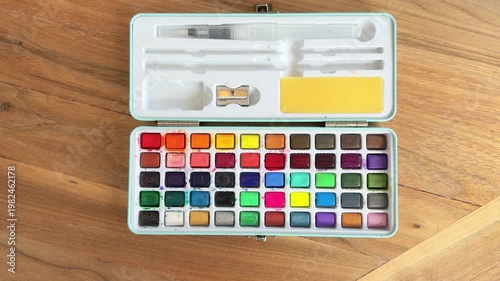 Top view of watercolor paint set being activated with water spray bottle, vibrant colors blending in palette. Close-up 4K art process on wooden table.