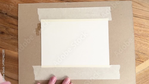Female Caucasian hands using a paintbrush to spread water across watercolor paper, preparing a wet surface for painting. Close-up 4K art technique on wooden table.