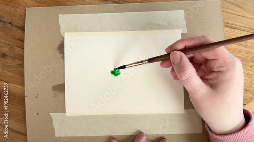 Female Caucasian hand dropping green watercolor pigment onto wet paper, paint blooming and spreading naturally. Close up 4K wet-on-wet painting technique on taped watercolor paper.
