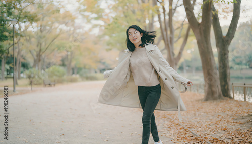 A woman joyfully walks through a tree-lined park path covered with autumn leaves, wearing a beige trench coat and casual outfit.