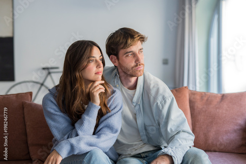 Принти на полотні Young couple sits together on a living room couch, sharing a quiet, thoughtful moment as they gaze out the window
