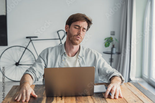 Canvastavla A young man sits at a wooden table with a laptop, eyes closed and head tilted ba