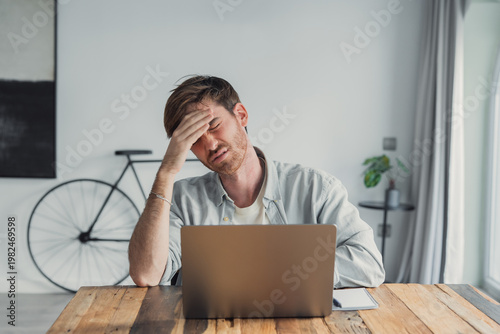 Foto Young man at a wooden table works from home on a laptop, holding his head in frustration