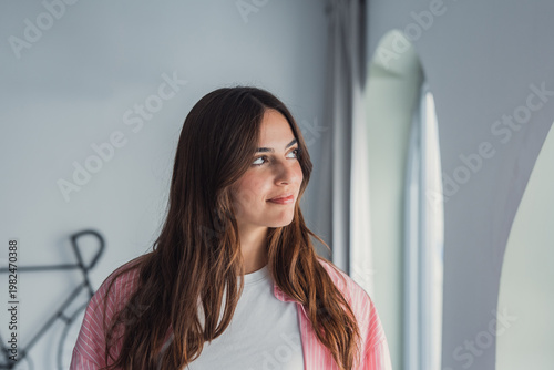 фотографія A young woman in a pink shirt gazes out a window in a bright modern interior, ap