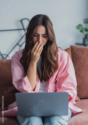 Young woman sits on a sofa using a laptop at home, covering her mouth in shock and worry while reading upsetting news, an emotional message or unexpected email online.