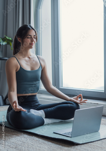 A young woman sits cross-legged on a mat by a bright window, meditating during an online yoga and mindfulness session for relaxation, fitness and mental wellness.