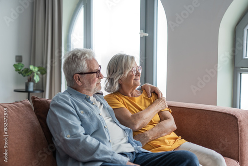 Older couple relax on a sofa in a bright living room, sharing a tender, affectionate moment. Warm, peaceful scene conveying love, companionship, retirement comfort and long-term partnership at home.