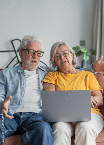 An older couple relaxes on their sofa while sharing a laptop, looking engaged as they browse, video chat, or learn online — capturing companionship, technology use, and everyday home life.