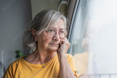 Fotografi A thoughtful senior woman in a yellow shirt looks out a window, appearing reflective and pensive