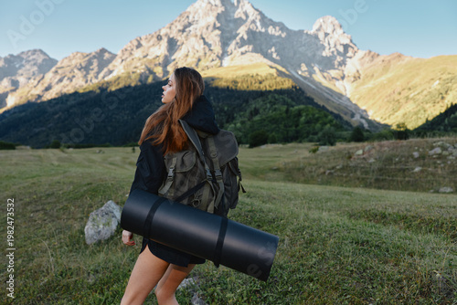 A lone hiker with a backpack and rolled mat traverses a grassy valley beneath rugged mountains. She gazes toward the horizon, ready for outdoor exploration.
