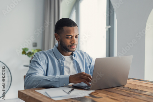 A young man concentrates while typing on a laptop at a wooden table in a bright home office, surrounded by papers and a pen, conveying remote work, concentration and everyday productivity.