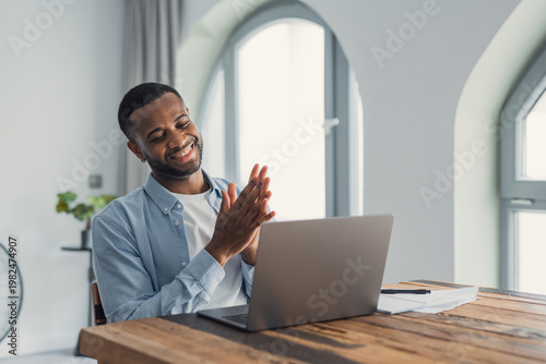 Vászonkép A smiling man applauds in front of his laptop, celebrating a successful remote work moment in a bright, modern home office