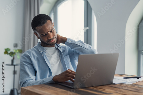 Young man at a wooden desk in a home office rubs his neck while working on a laptop, showing discomfort from long screen time, poor posture and remote work strain. Documents and pen nearby.