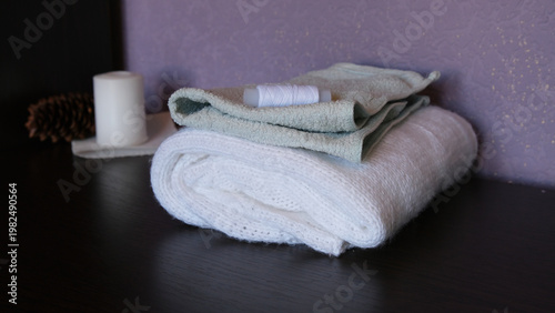 A spool of white thread lies on top of light green and white folded terry towels on a dark wooden table next to a white candle.