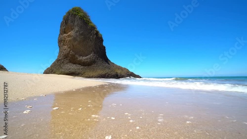 Low angle view of clear ocean waves washing onto a golden sandy beach with a prominent rock formation under a vibrant blue sky.
