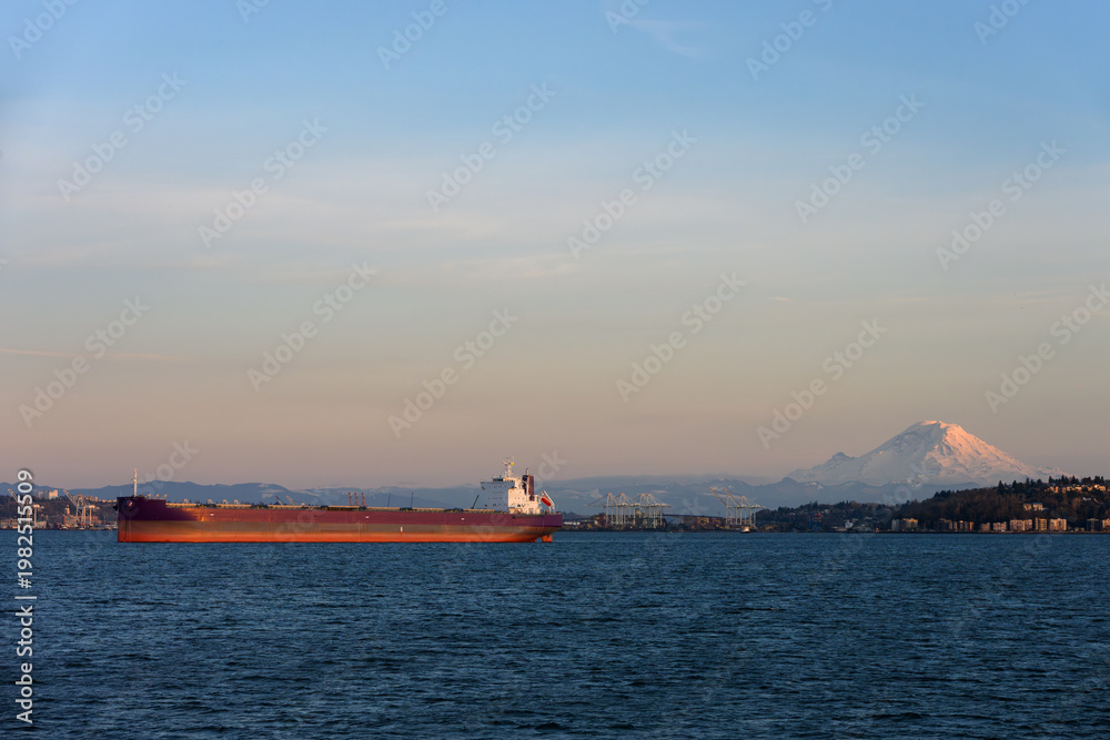 Fototapeta premium Large cargo ship transporting goods in Puget Sound at sunset, international freight, trade, commerce, and economics in action, Mt. Rainier and port of Seattle in background 