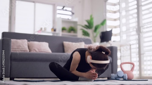 Asian adult woman perform home workout stretching exercise on yoga mat with headphones in modern living room promoting healthy lifestyle wellness flexibility