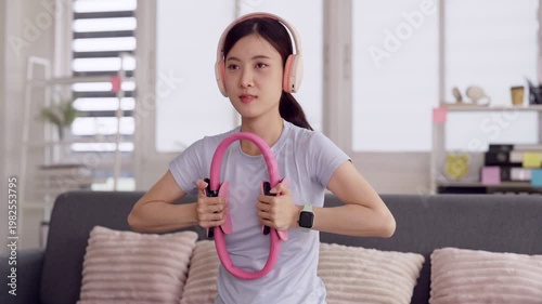 Asian young adult woman exercising pilates ring home living room wearing headphones smartwatch for fitness wellness health workout routine