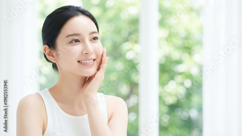 やわらかな自然光が差し込む窓辺で頬に手を添えて微笑む若い女性のナチュラルビューティーポートレート、素肌感や透明感、保湿ケアを印象づけるスキンケア広告向け人物素材
