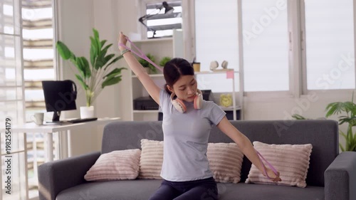 Adult Asian woman home workout resistance band exercise stretching fitness sitting sofa modern apartment living room active lifestyle wellness healthy routine