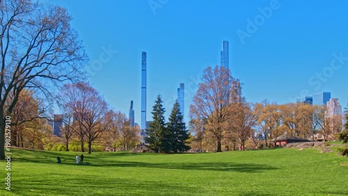 A static 4K wide-angle shot of Central Park's green meadow on a bright spring day. Unrecognizable people relax on the lawn while the towering skyscrapers are visible in the background