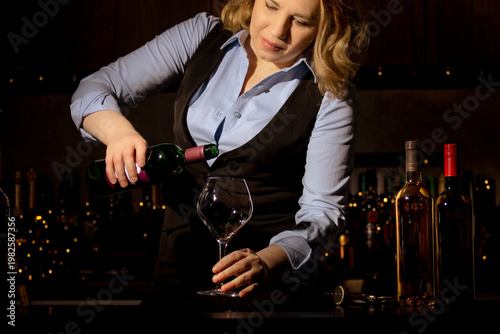 Professional woman sommelier pouring red wine into glass at bar counter. Elegant beverage presentation and precise restaurant service technique.