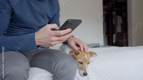 Man using smartphone while sitting on bed with small dog in home bedroom. Concept of mobile communication pet companionship home lifestyle and everyday technology