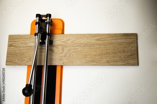 A top view of an orange mechanical tile cutter with a metal river, on which ceramic tiles of wooden colors lie sideways on a white background. The concept of renovation, tiling, construction work