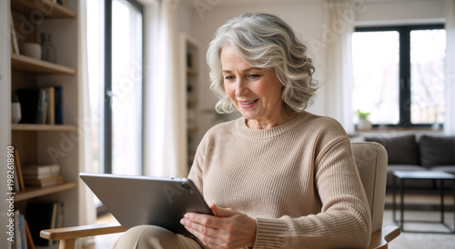 Senior woman using a digital tablet in a modern living room. Smiling elderly female looking at screen at home. Technology and communication concept