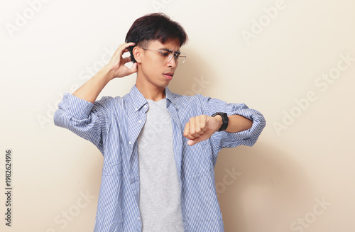 Photography Confused young Asian man looking at wristwatch while scratching head with stressed gesture on isolated background
