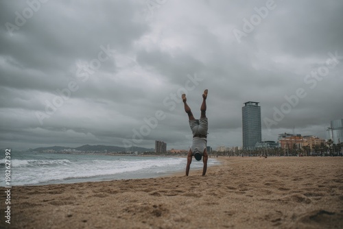 A solitary figure performs a handstand on a sandy beach under a cloudy sky, with the cityscape visible in the background, capturing a moment of athleticism and coastal serenity.