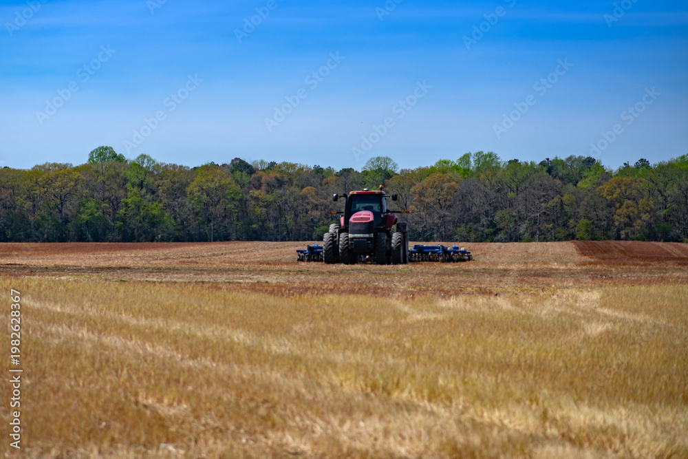 Fototapeta premium Tractor operates on plowed field soil. Tractor supports heavy tillage with disc. Farm machinery uses tractor for cultivation. Blue sky frames tractor in landscape.