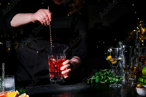 Woman bartender with bar spoon and red Manhattan cocktail in mixing glass. Professional mixology and drink dilution control.