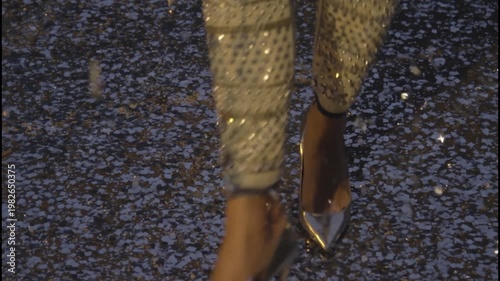 A woman's legs wearing metallic high heels and sequined pants on a glowing asphalt road at night.

