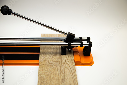 Side view of an orange mechanical tile cutter with a metal river, on which ceramic tiles of wooden color lie sideways on a white background on the side with space for text. The concept of renovation