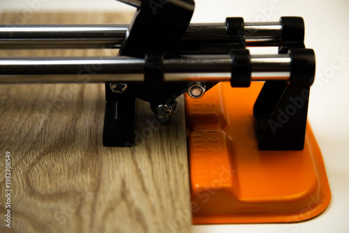 Close-up side view of an orange mechanical tile cutter, which fixed ceramic tiles of wooden colors, above it is a cutting disc on a white background. The concept of renovation, tiling, construction