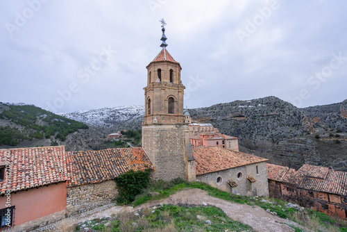 A scenic view of the historic medieval town of Albarracin in Spain, featuring stone buildings, a church tower, and snow-capped mountains in the background.