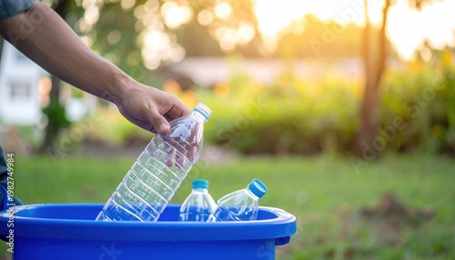 Hand throwing plastic bottle into recycling bin outdoors at sunset.