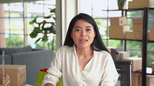 Woman business owner greeting audience on camera while working in home office with packaging boxes, ideal for online business and content creation
