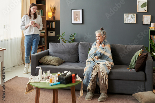 Картината върху платно Senior Caucasian woman sitting wrapped in blanket while young adult daughter holding mug watching over her at home, showing illness care and family support