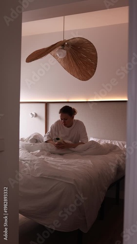 Young woman lying in white bed scrolling smartphone in cozy stylish bedroom with warm rattan pendant lamp. Perfect for lifestyle, technology, relaxation and evening routine themed projects