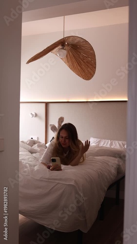 Young woman lying in white bed having video call on smartphone in stylish bedroom with rattan pendant lamp. Perfect for lifestyle, technology, communication and morning themed projects