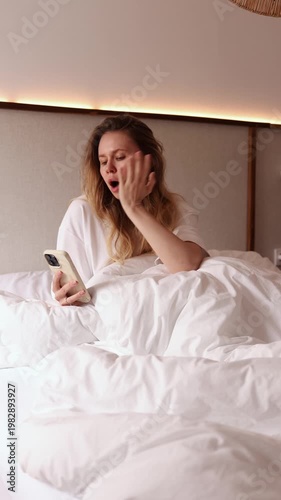 Sleepy young woman yawning and scrolling smartphone while sitting in white bed in the morning. Perfect for lifestyle, morning routine, technology and relatable emotion themed projects