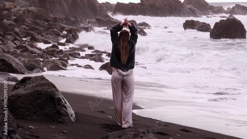 Young woman raising arms up in freedom on dramatic black volcanic sand beach with powerful Atlantic ocean waves and rocks in Tenerife. Perfect for travel, lifestyle and emotion projects